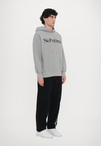Grauer Kapuzenpullover mit "No Problemo" in schwarzer Schrift, kombiniert mit schwarzen Hosen. Der Hoodie hat eine Kordel mit einem kontrastierenden Muster.