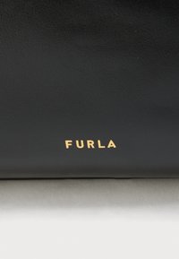 Must tekstureeritud nahkpind kuldse suure algustähega "FURLA" brändinimega, mis asub peaaegu põhjaserva keskel.