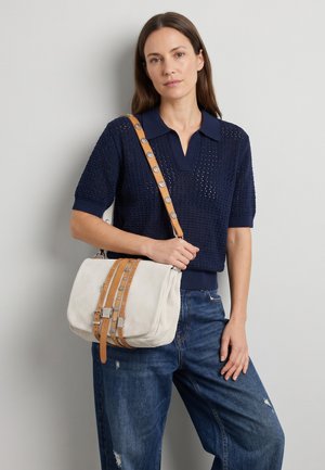 Vrouw draagt een marineblauwe gebreide top met korte mouwen en blauwe spijkerbroek, houdt een witte schoudertas met beige riemen en metalen studs vast.
