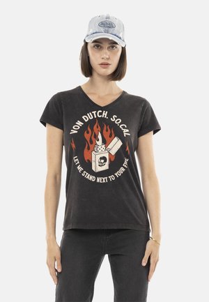 Jeune femme aux cheveux bruns raides porte une casquette bleu clair, un T-shirt noir Von Dutch avec un motif de flammes et de briquet, et un jean noir.