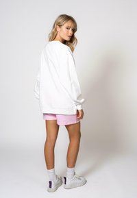 Sweatshirt blanc à coupe oversized, associé à un short rose clair. Le modèle porte des baskets blanches avec des accents violets et des chaussettes blanches.
