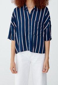 Chemise bleu marine à manches courtes avec rayures verticales blanches et rouges, poche poitrine unique, coupe décontractée, tissu léger. Pantalon blanc.