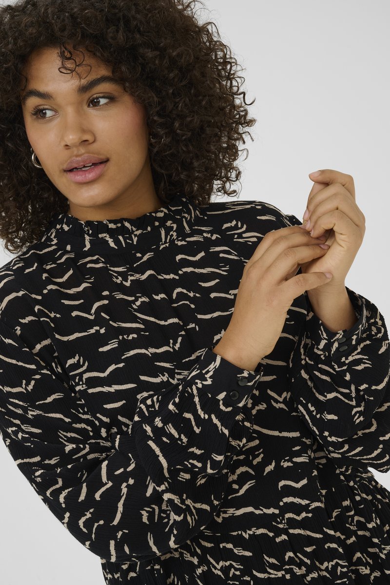 Blouse noire à motifs avec un col volanté, ornée de lignes abstraites crème. Manches longues avec poignets boutonnés. Tissu texturé.