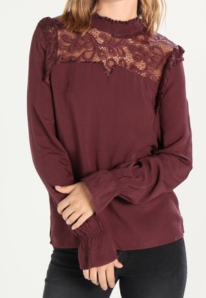 Kvinde iført en langærmet vinrød bluse med blonder på skuldre og øverste del af brystet, kombineret med sorte bukser.