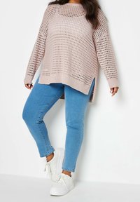 Pull en tricot rose clair avec une coupe ample, un motif en maille ajourée, des manches longues, fentes sur les côtés, assorti à un jean slim bleu et des baskets blanches.