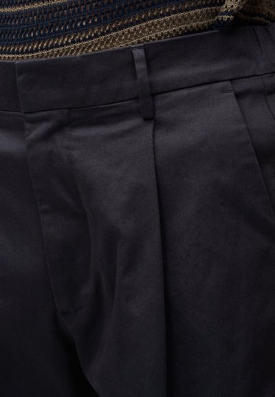 Gros plan d'un pantalon noir sur mesure avec passants de ceinture et plis, porté avec un top en maille marron et bleu.