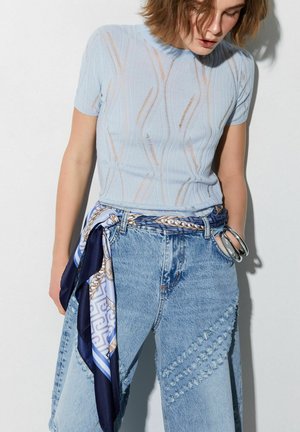 Donna che indossa una maglietta a maniche corte di colore azzurro chiaro con fantasia, jeans a vita alta con toppe e una sciarpa fantasia utilizzata come cintura.