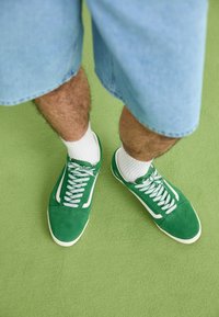 Personne portant des baskets vertes et blanches, des chaussettes blanches côtelées et un short en jean bleu clair debout sur un tapis vert.