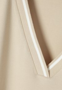 Top beige avec un design col en V. L'encolure présente des bandes blanches en accent et un fini en tissu lisse et texturé.