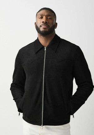 Man with short hair and beard wearing a black zip-up jacket with hands in pockets against a plain light background.