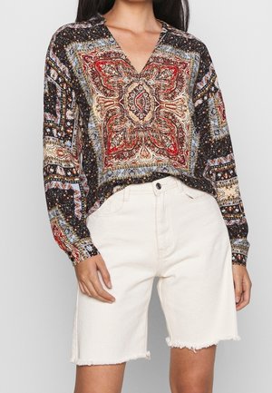 Vrouw draagt een blouse met lange mouwen met een patroon in rood en blauw en een witte denimshort met hoge taille en rafelige zomen.