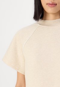 T-shirt beige a maniche corte con colletto rotondo; presenta cuciture decorative lungo le spalle e una leggera texture. Materiale morbido simile al cotone.