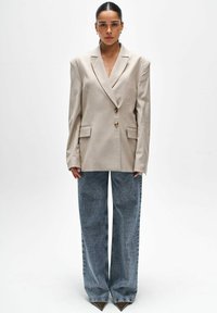 Beige oversized blazer met notch-lapels, dubbelknopen van goud en voorzakken; gecombineerd met lichtblauwe wijde jeans en puntige hakken.