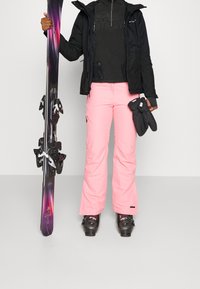 Skidkläder med en svart isolerad jacka, svart dragkedjeskjorta, rosa vattentäta skidor, och svarta pjäxor, som håller i flerfärgade skidor.