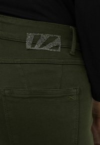 Jeans verde oliva con un parche de logo plateado brillante en la parte posterior de la cintura y detalles de costura mínimos. Presenta un diseño clásico de cinco bolsillos.