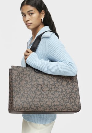 Kobieta w jasnoniebieskim swetrze niesie dużą torbę w brązowy wzór z czarnymi uchwytami na ramieniu na jednolitym tle.