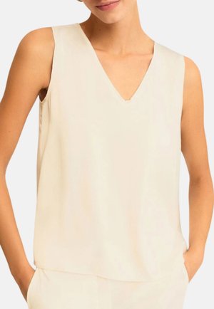 Donna che indossa un top senza maniche color crema con scollo a V e le mani nelle tasche di pantaloni crema coordinati su sfondo bianco.