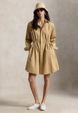 Kvinde iført en beige skjortekjole med snor i taljen, bredskygget hat, sorte solbriller, hvide sneakers og perledråbeøreringe.