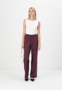Bílý žebrovaný top bez rukávů v kombinaci s vysokými, širokými kalhotami v barvě maroon. Modelka drží malou bílou tašku a má na sobě černé špičaté boty.