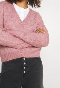 Osoba v růžovém pleteném kardiganu s texturou přes bílou košili s límečkem, se zkříženýma rukama a v černých džínách s vysokým pasem a knoflíkovým zapínáním.