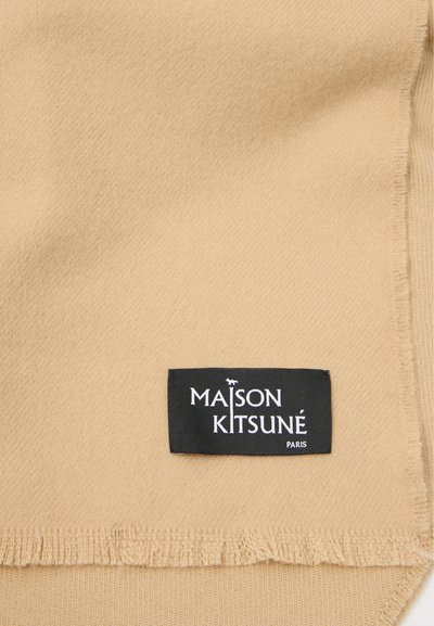Μπεζ ύφασμα δείγμα με μαλακή υφή, φέρει μια μαύρη ετικέτα που γράφει "MAISON KITSUNÉ PARIS" με λευκά γράμματα. Οι άκρες είναι φραγμένες.