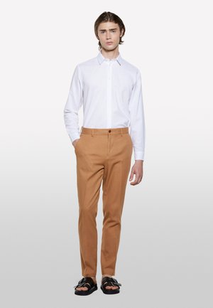 Camicia bianca con bottoni, pantaloni tailored beige, tessuto strutturato, e sandali neri con dettaglio a fibbia. Design semplice e moderno con una vestibilità slim.