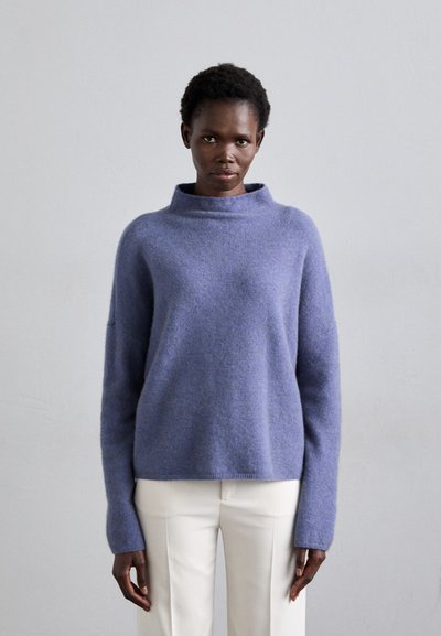 Pull en laine violet avec un col montant, des manches longues et une coupe décontractée. Tissu léger, associé à un pantalon couleur crème.