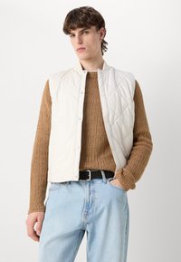 Vesta albă matlasată cu guler înalt, închidere cu capse și design fără mâneci, purtată peste un pulover texturat maro și blugi de culoare bleu deschis.