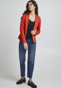 Rode blazer met een figure-hugging ontwerp, gecombineerd met een zwarte top met kantafwerking en blauwe rechte jeans. Zwarte loafers met gouden accenten.