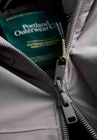 Siva vanjska jakna s crnom unutarnjom postavom, koja se ističe čvrstim patentnim zatvaračem sa žutim akcentima. Oznaka kaže "Portland Outerwear Co."