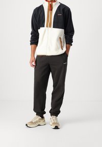 Giacca in pile con collo alto in blu scuro e crema, con un pannello marrone; abbinata a pantaloni jogger scuri e scarpe da ginnastica beige.