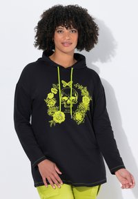 Černá mikina s kapucí, která má neonově zelený květinový skull grafický design, tkaničky a kontrastní švy. Nositelná přes zelené kalhoty.