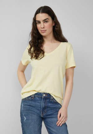 Jonge vrouw met lang donker krullend haar, die een lichtgeel V-hals T-shirt en een blauwe spijkerbroek draagt, staand tegen een effen grijze achtergrond.