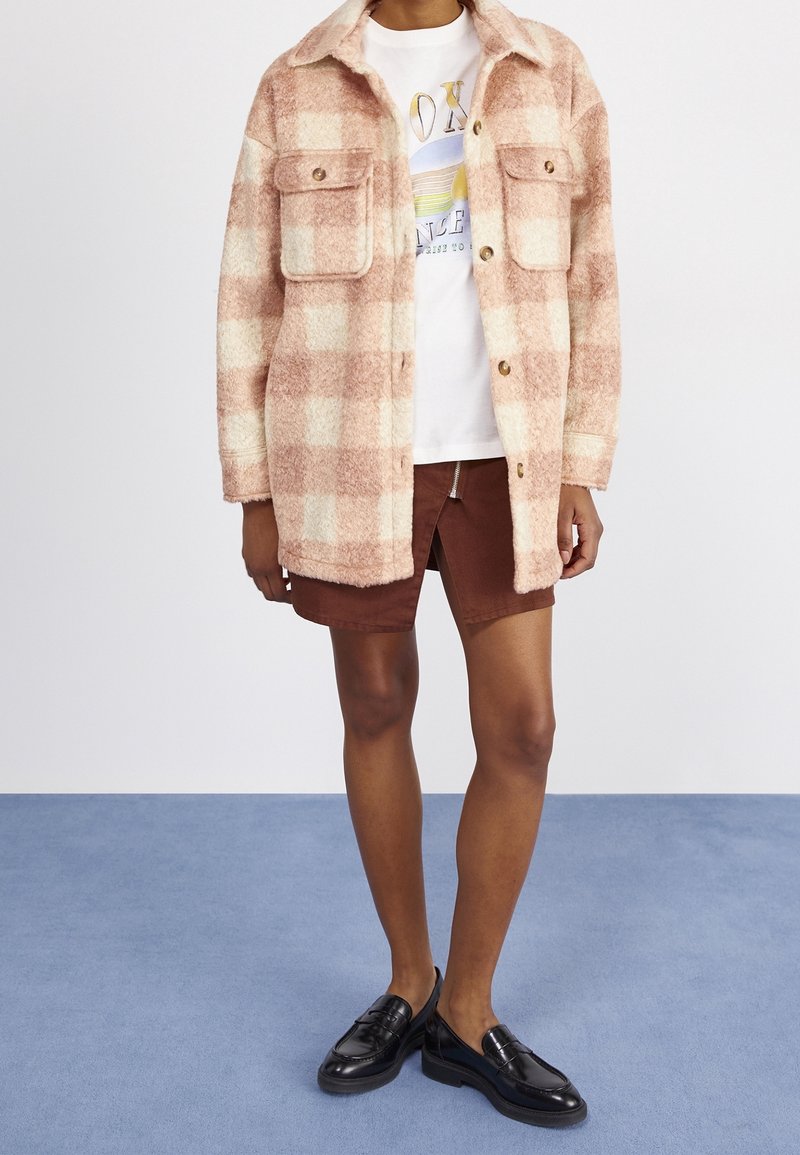 Personne portant une longue veste à carreaux rose et crème sur un T-shirt blanc à motif, une jupe marron fendue à l'avant et des mocassins noirs.