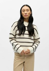 Kreminės spalvos megztinis su rudo atspalvio horizontaliomis juostelėmis, storu mezgimo raštu, su riboto stiliaus rankogaliais ir apvalia iškirpte, dėvimas ant šviesiai smėlio spalvos kelnių.