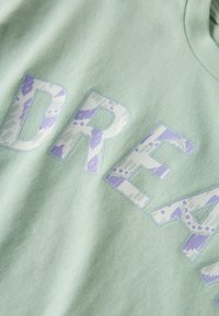 T-shirt en coton vert clair avec le texte "DREAM" en grand motif blanc et lavande, au toucher doux et au design col rond.