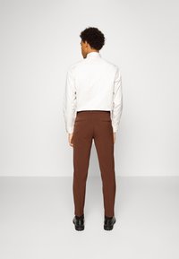 Pantaloni marroni su misura realizzati in tessuto morbido, con una vestibilità slim, tasche laterali e una vita senza cuciture. Abbinati a una camicia di colore chiaro.