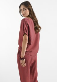 Burgundský outfit s texturovaným topem s krátkým rukávem a odpovídajícími zúženými kalhotami, doplněný širokým tmavě červeným náramkem.