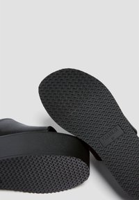 Sandales à plateforme noires avec une tige lisse et une semelle en caoutchouc texturée présentant un motif ondulé. Nom de la marque imprimé sur la semelle.