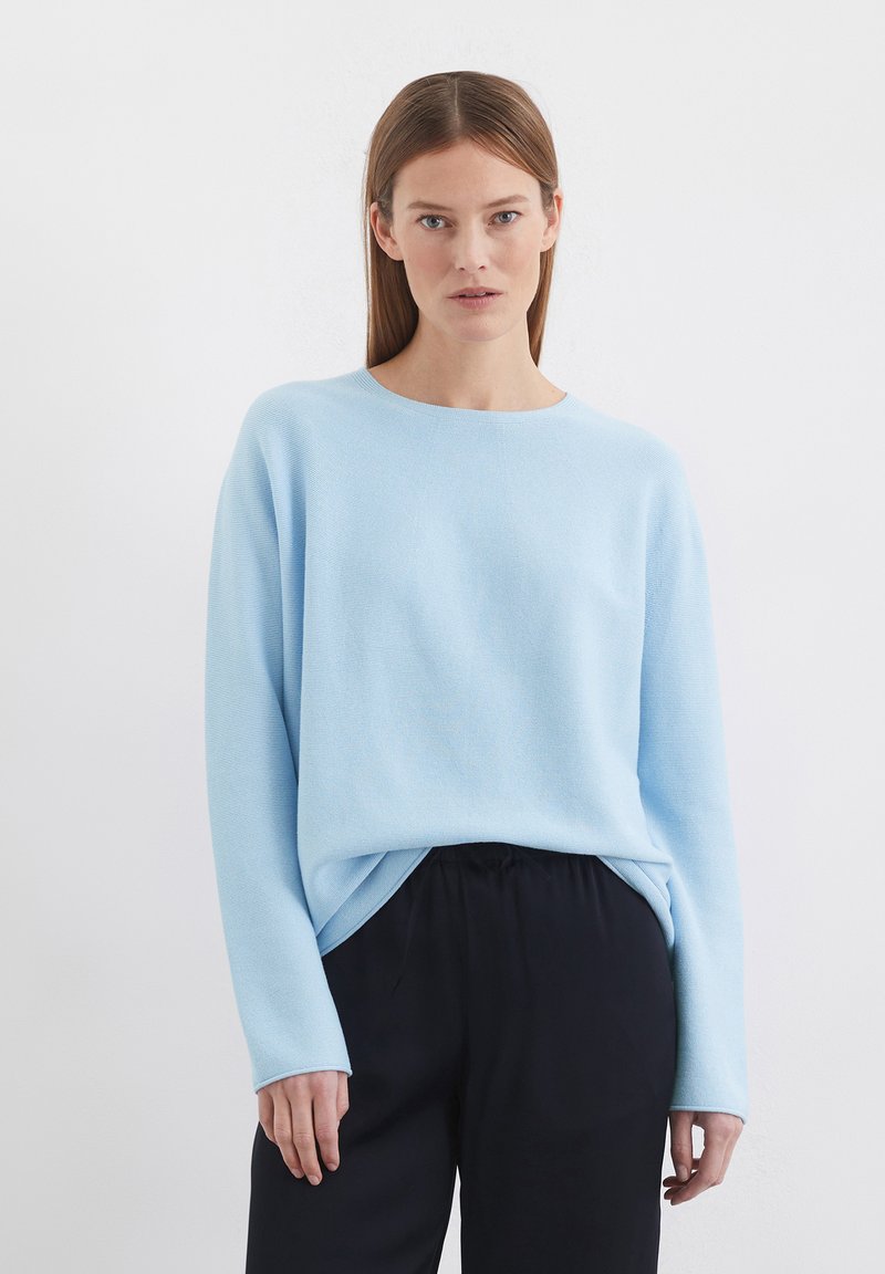 Pull en tricot bleu clair avec un col rond, des manches longues et une coupe décontractée, associé à un pantalon noir. Texture lisse, design décontracté.
