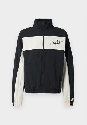 Sort og cremefarvet vindtæt jakke med høj krave, fuld lynlås, elastiske manchetter og broderet Nike-logo på brystet. Tekstureret stof.