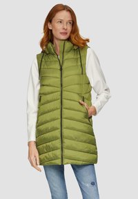 Grüne, gepolsterte Weste mit hohem Kragen, vertikalen Nähten, zwei Seitentaschen und einem durchgehenden Reißverschluss. Getragen über einem weißen Langarmoberteil.