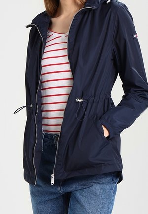 Femme porte une veste zippée bleu marine sur une chemise blanche rayée rouge, main dans la poche de la veste, associée à un jean bleu.