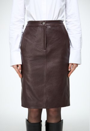 Žena nosi hnedú koženú sukňu po kolená, bielu košeľu s dlhým rukávom, priehľadné čierne pančuchy a čierne čižmy po kolená.