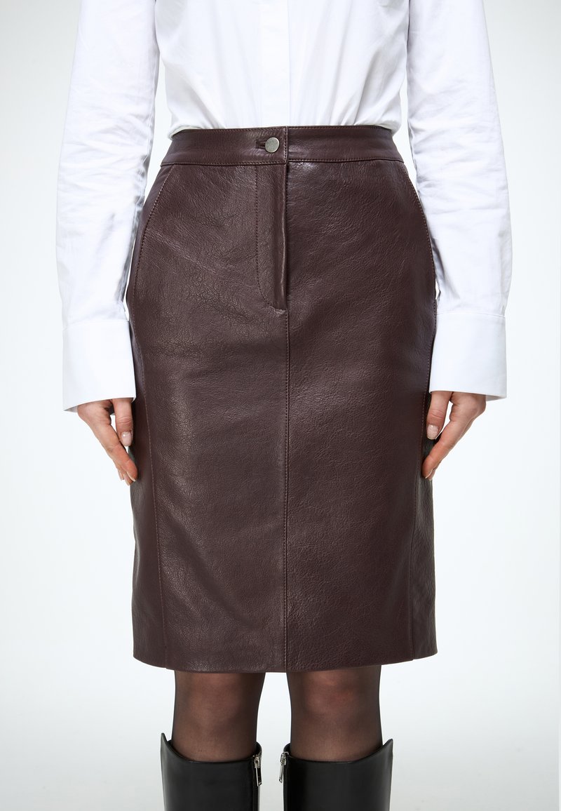 Žena nosi hnedú koženú sukňu po kolená, bielu košeľu s dlhým rukávom, priehľadné čierne pančuchy a čierne čižmy po kolená.