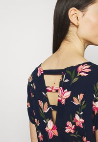 Laivaston sininen kukkamekko, jossa on vaaleanpunaisia ja valkoisia kukkia, pieni aukko takana ja lyhyet hihat rentoa istuvuutta varten.