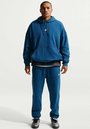 Mies seisoo päällään sininen Nike-huppari ja yhteensopivat verkkarit, kädet sivuilla, katsoo eteenpäin, harmaat lenkkarit jalassa, yksivärisellä taustalla.