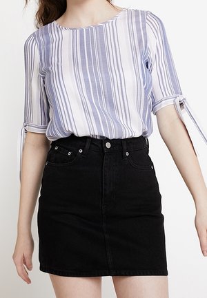 Femme portant un chemisier à rayures verticales bleu et blanc avec des détails de nouage sur les manches et une jupe en denim noire taille haute.