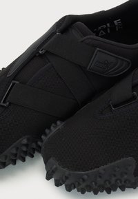 Zapatillas deportivas negras con material de malla, diseño de correas elásticas, suela de goma texturizada con picos salientes y detalles de marca.