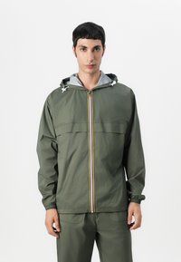 Giacca leggera di colore verde oliva con zip frontale, fodera della cappuccio grigia, polsini elastici e una striscia verticale colorata al centro.
